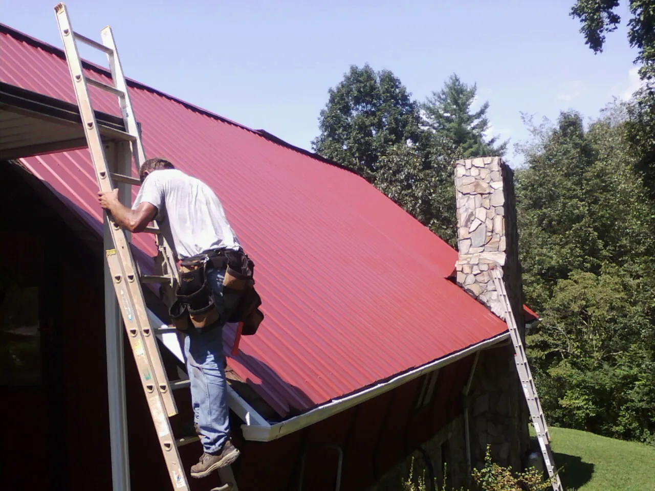 Roofer on a ladder beside a red metal roof and stone chimney — Soffit & Fascia Repair by Roofing Brothers in Caddo Mills